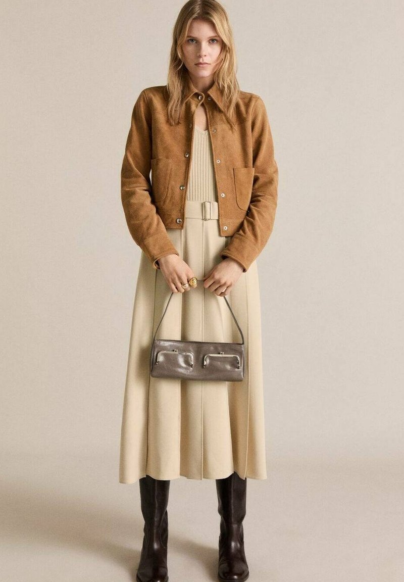 Giacca in suede marrone, abito plissettato beige, stivali marroni fino al ginocchio e borsa argentata. Texture morbide, silhouette classica e design semplice.