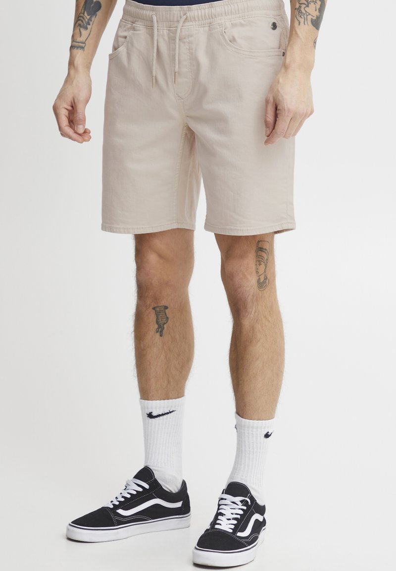 Beige shorts with an elastic waistband, drawstring, and side pockets. Worn with black sneakers and white socks featuring a logo.