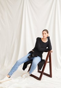 Chemise noire à manches longues, jean skinny bleu clair et baskets blanches. Modèle assis sur une chaise en bois avec une veste sombre drapée dessus.