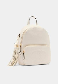 Zaino in finta pelle color crema con accessori dorati, tasca frontale con zip e lungo dettaglio frangiato. Forma arrotondata e finitura testurizzata.