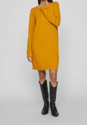 Mustard yellow knitted dress with long sleeves and V-neck, featuring ribbed cuffs and hem, paired with black knee-high boots.