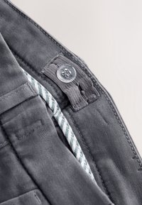 Gros plan de la ceinture d’un pantalon gris montrant une patte de bouton réglable et une bande élastique rayée à l’intérieur.