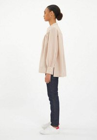 Blouse rayée à manches longues en beige clair et rose, présente un col haut, des manches ballons et des poignets boutonnés. Associée avec un jean foncé et des baskets.