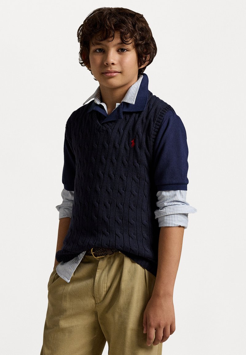 Σκούρο μπλε πλεκτό γιλέκο με καπιτονέ σχέδιο πάνω από ανοιχτό μπλε πουκάμισο με ρίγες και γιακά, συνδυασμένο με κρέμ παντελόνι και καφέ ζώνη.