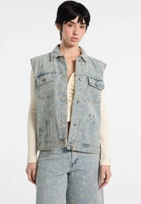 Jasnoniebieska dżinsowa kamizelka bez rękawów z przednimi kieszeniami, zniszczoną fakturą i srebrnymi zatrzaskami, noszona na kremowej bluzce z długim rękawem.