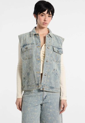 Giacca senza maniche in denim azzurro chiaro con tasche anteriori, texture strappata e bottoni a pressione argentati, indossata sopra una maglia a maniche lunghe crema.