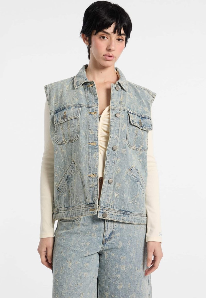 Jasnoniebieska dżinsowa kamizelka bez rękawów z przednimi kieszeniami, zniszczoną fakturą i srebrnymi zatrzaskami, noszona na kremowej bluzce z długim rękawem.