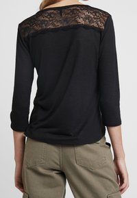 Svart topp med trekvartsärmar, med en spetspanel på baksidan och vågformad trim. Mjuk textur, avslappnad design.