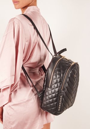 Mochila negra acolchada con cremalleras doradas, con correas ajustables y detalles laterales, llevada sobre una prenda de color rosa claro.