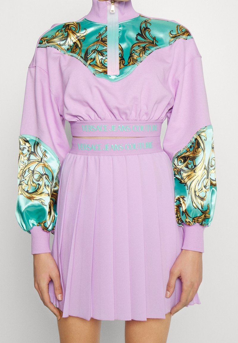 Lila klänning med hög krage och dragkedja, med turkosa satinärmar prydda med gyllene mönster och en plisserad kjol.