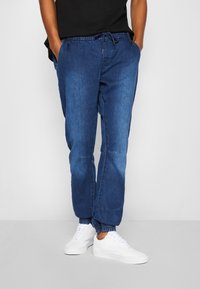 Joggers de mezclilla en azul oscuro con cintura elástica y puños. Incluyen bolsillos laterales y un cordón para un ajuste ajustable. Combinados con zapatillas blancas.