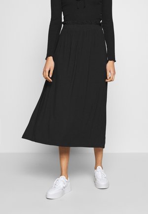 Kvinde iført sort langærmet top og sort midi nederdel med indsnævret talje, kombineret med hvide plateau sneakers, stående op ad en grå væg.