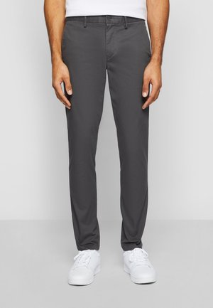 Mand iført mørkegrå slim-fit bukser og hvide sneakers, står foran en ensfarvet lys baggrund.