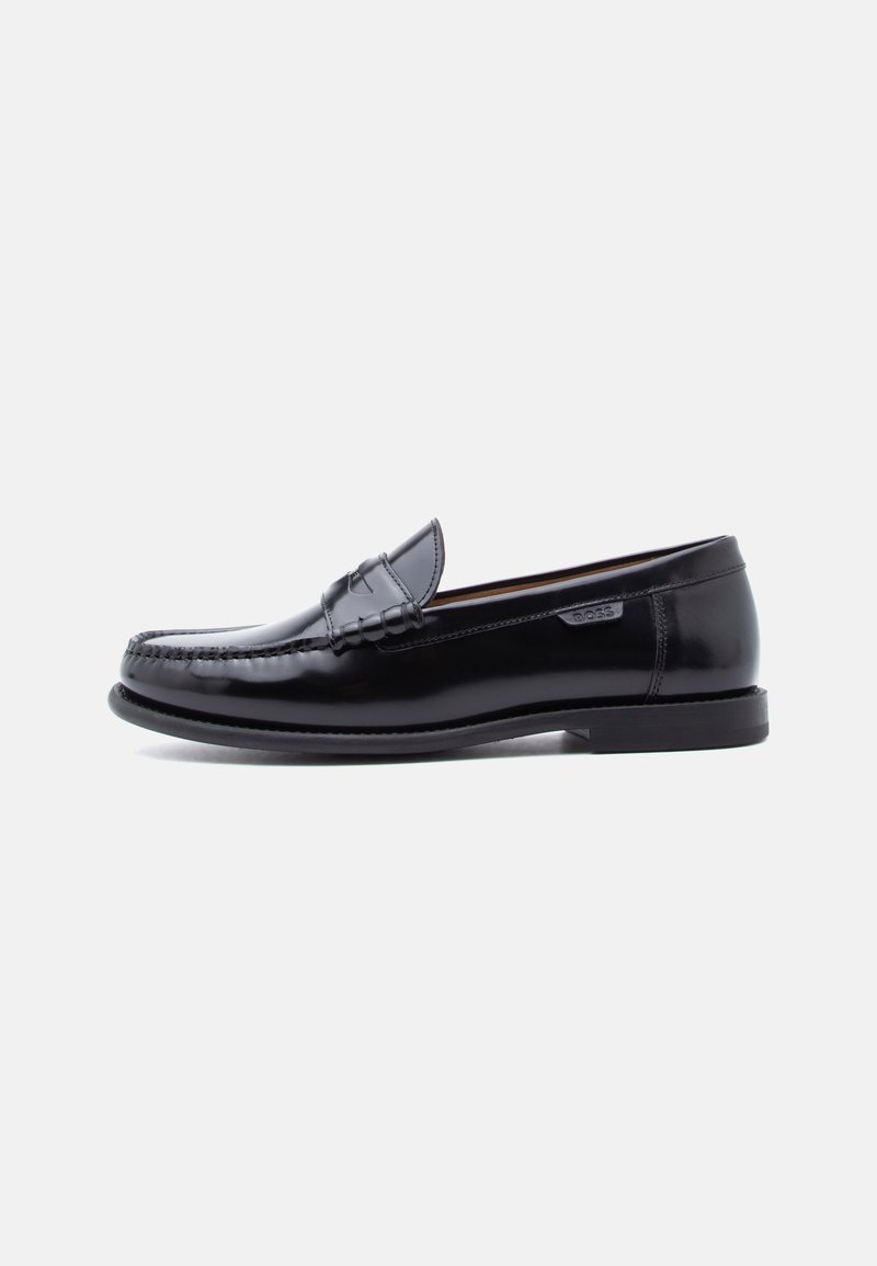 Mocassins en cuir noir avec une finition brillante, présentant un motif cousu sur l'avant-pied et une semelle en caoutchouc noire texturée et basse.