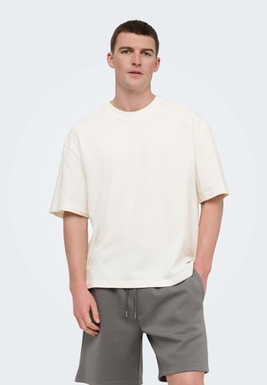 Ung mand med kort hår iført en løs hvid T-shirt med korte ærmer og grå shorts med snøre, stående med den ene hånd i lommen.