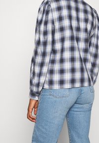Camicia a scacchi blu e bianca con maniche a sbuffo, realizzata in un tessuto leggero, abbinata a jeans blu chiaro. Chiusura con bottoni visibile ai polsini.