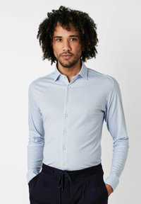 Hellblaues Langarmhemd mit geometrischem Muster, Button-down-Kragen und weißen Knöpfen. Der Stoff hat eine glatte Textur. Kombiniert mit dunklen Hosen.