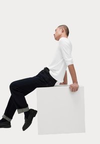 Weißes Langarmhemd, dunkelblaue Denim-Jeans mit hochgekrempelten Säumen und schwarze Sneaker. Auf einem weißen Würfel sitzend, seitlich posiert.