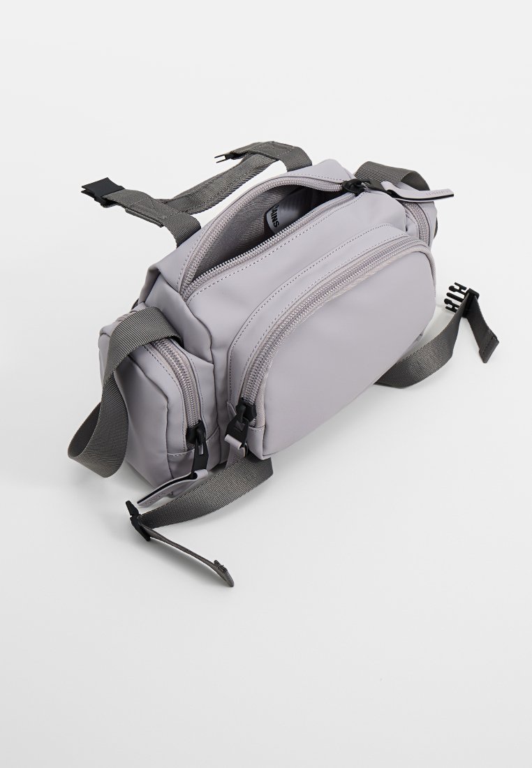 Lichtgrijze stoffen heuptas met drie ritscompartimenten en verstelbare donkergrijze banden op een witte achtergrond.