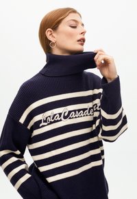 Námořnický svetr s krémovými pruhy, s vysokým límcem a vyšitým detailem "Lola Casademunt". Hřejivá struktura a oversized fit.