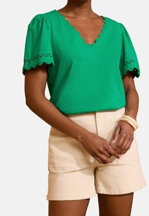 Kobieta w zielonej bluzce z krótkim rękawem z dekoltem w serek z falbanką i beżowych szortach z wysokim stanem, na lewym nadgarstku ma dwie kremowe bransoletki.