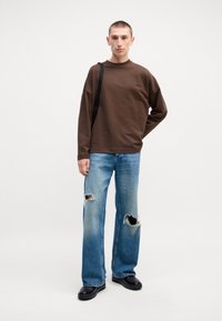 Chemise marron à manches longues, coupe décontractée. Jean bleu clair déchiré avec des bords effilochés. Chaussures noires, debout sur un fond neutre.