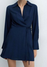 Námořnická modrá zavinovací šaty s dlouhými rukávy a límcem. Vyrobeno z lehké látky, má pásek v oblasti pasu a sukně je rovného střihu.