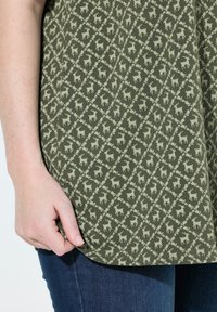 Zelená látka se vzorem obsahuje světlé motivy jelenů uspořádané do diamantového designu. Ruka je umístěna na lemu, což doplňuje džíny.