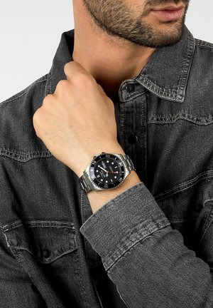 Edelstahluhr mit schwarzem Zifferblatt, silbernen Markierungen und roten Akzenten, getragen an einer Hand vor einem dunklen Denimhemd mit Knopfdetails.