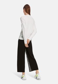 Hvid langærmet skjorte med et logo på ryggen, kombineret med sorte bukser med vide ben, der har hvide geometriske detaljer langs siderne.