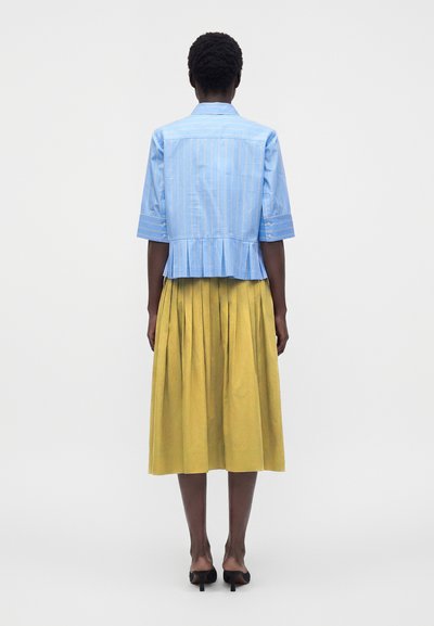 Femme debout de dos portant une chemise plissée bleu clair à manches courtes et une jupe midi plissée jaune moutarde avec des talons noirs.