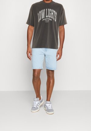 T-shirt oversize grigio scuro con testo grafico bianco, pantaloncini di jeans azzurri, e sneakers grigie con lacci bianchi e dettagli del logo.