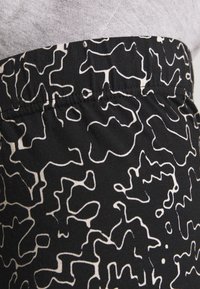 Černá tkanina s bílým abstraktním vzorem. Obsahuje elastický pás a měkký, texturovaný povrch. Detailní záběr na detaily oděvu.