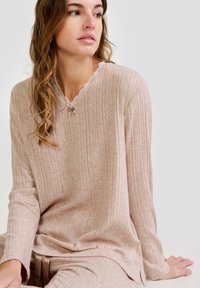 Mujer con cabello ondulado vestida con ropa de lounge de punto acanalado beige, con un escote en V adornado con encaje y un pequeño lazo, sentada frente a un fondo sencillo.