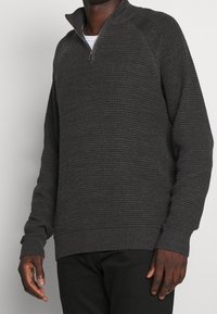 Mann trägt einen dunkelgrauen, strukturierten Sweatshirt mit Viertelreißverschluss und schwarze Hosen, steht vor einem hellen, einfarbigen Hintergrund.