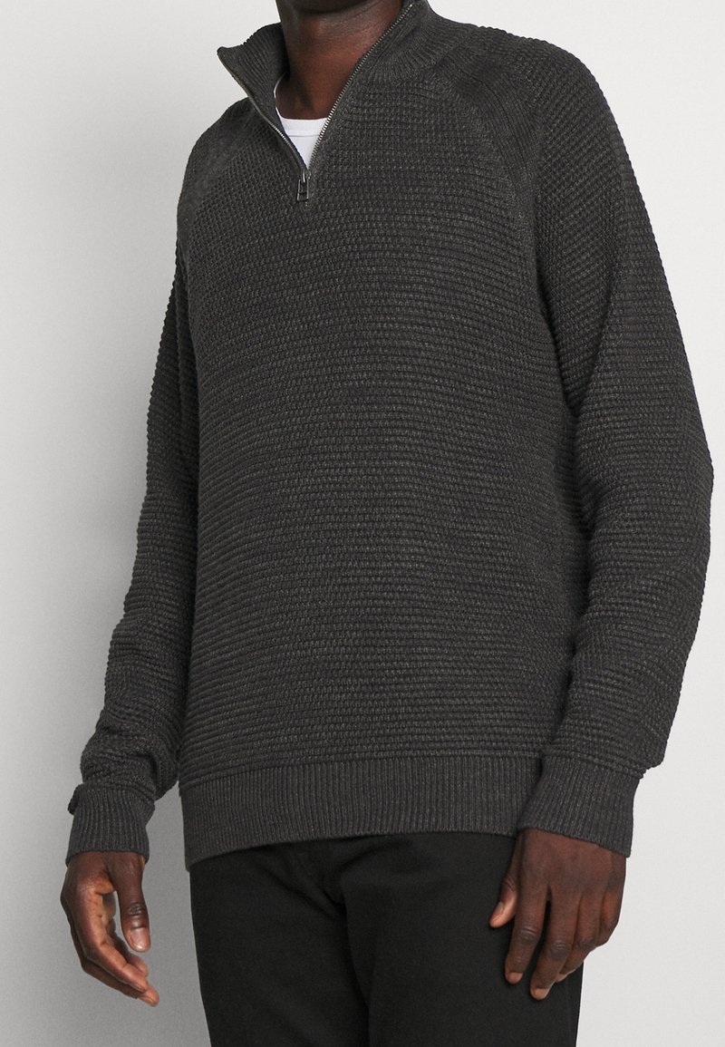 Mann trägt einen dunkelgrauen, strukturierten Sweatshirt mit Viertelreißverschluss und schwarze Hosen, steht vor einem hellen, einfarbigen Hintergrund.