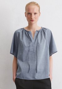 Blusa di cotone leggera, a maniche corte, di colore azzurro con scollo a V, dettaglio arricciato sul colletto e vestibilità rilassata. Indossata con pantaloni scuri.