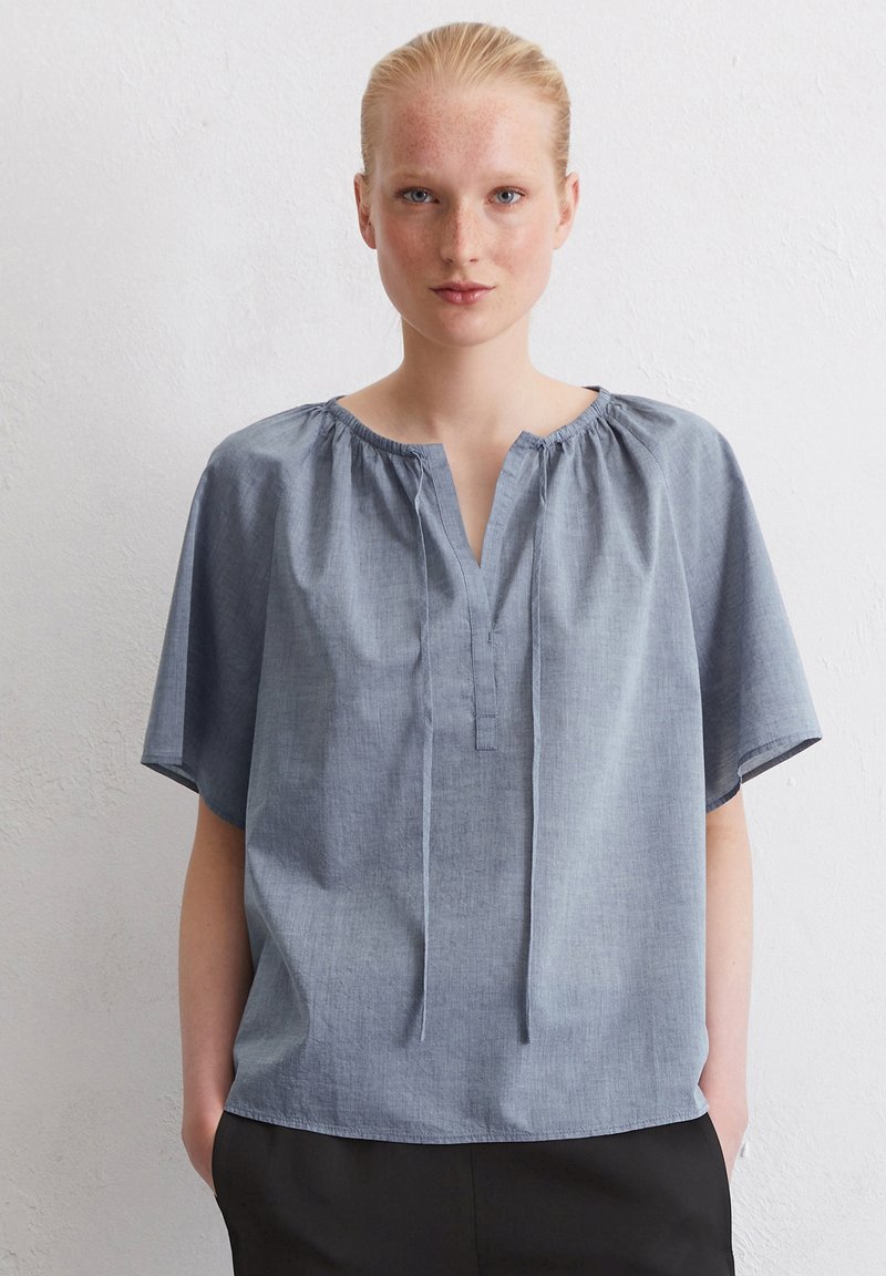 Blusa di cotone leggera, a maniche corte, di colore azzurro con scollo a V, dettaglio arricciato sul colletto e vestibilità rilassata. Indossata con pantaloni scuri.