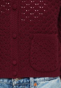 Bourgondische gebreide cardigan met een gestructureerd patroon, voorzien van ronde knopen, een patchzak en een geribde zoom. Gedragen over lichtblauwe jeans.