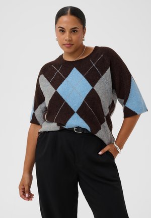 Femme aux cheveux foncés portant un pull à losanges marron, gris et bleu et un pantalon noir, posant avec une main dans la poche devant un fond uni.
