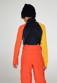 Ski-outfit met een meerkleurig shirt met lange mouwen in oranje, marineblauw en geel, met grijze accenten, gecombineerd met feloranje broeken en zwarte bretels.