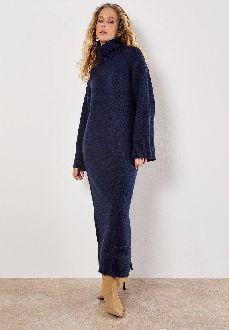 Vestito a collo alto lavorato a maglia di colore navy con maniche lunghe e spacco laterale, abbinato a stivaletti beige alla caviglia, su uno sfondo neutro.