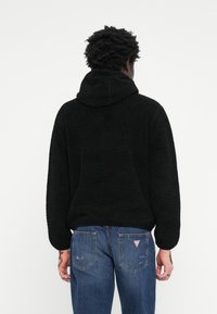 Svart fleece hettegenser med teksturert overflate, justerbar hette og løs passform. Kombinert med klassiske blå denimjeans med baklommer.