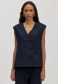 Vrouw in een marineblauwe mouwloze dubbelzijdige blazer met zes zwarte knopen en bijpassende marineblauwe broek, staand tegen een effen achtergrond.