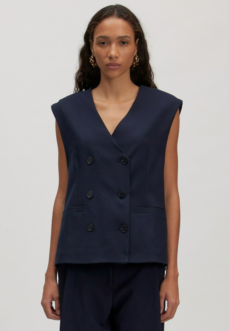 Vrouw in een marineblauwe mouwloze dubbelzijdige blazer met zes zwarte knopen en bijpassende marineblauwe broek, staand tegen een effen achtergrond.
