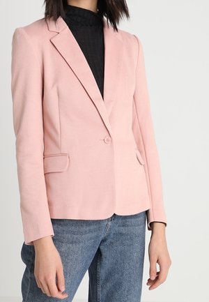 Woman wearing a fitted light pink blazer over a black top and blue jeans, standing against a plain light gray background.