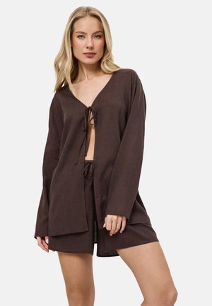 Femme blonde portant un haut noué à manches longues marron foncé et un short assorti, debout avec une expression neutre sur un fond clair.