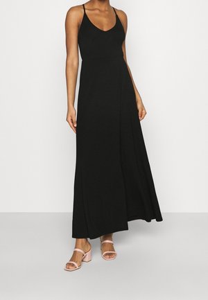 Žena v černých dlouhých šatech bez rukávů s tenkými ramínky a růžových sandálech na podpatku stojí před jednoduchým bílým pozadím.