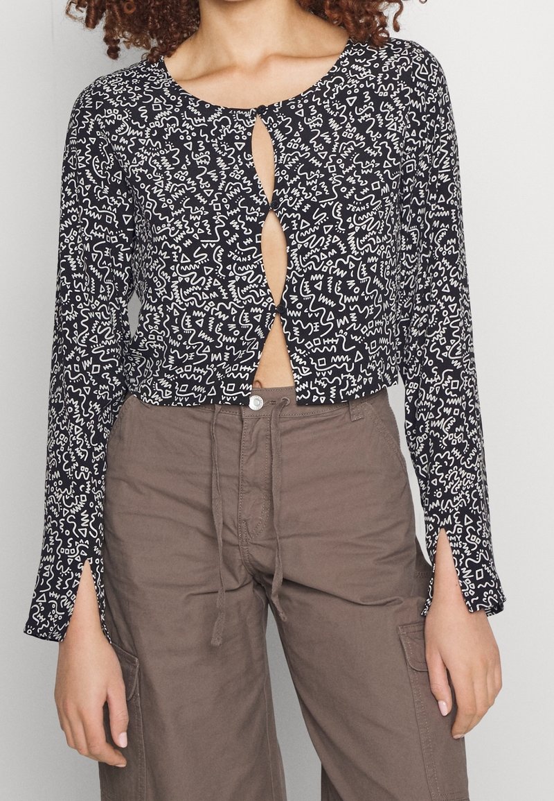 Femme portant un top court à manches longues boutonné avec un motif noir et blanc, et un pantalon cargo marron taille haute.