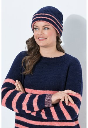 Frau trägt marineblauen und rosa gestreiften Strickhut und passenden Pullover, Arme verschränkt, lächelnd und zur Seite schauend.